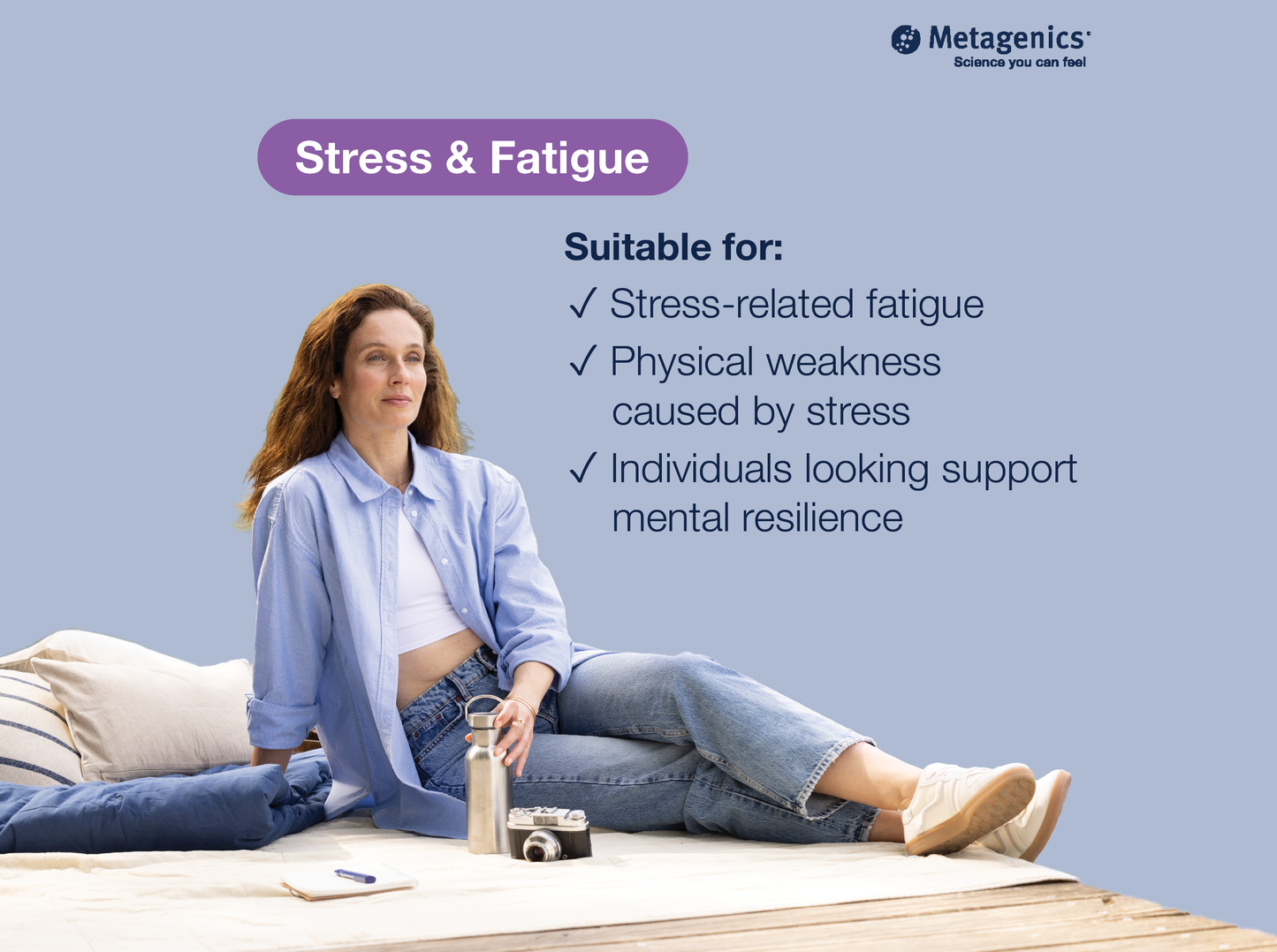 A woman sits on a wooden floor with pillows, wearing an open light blue shirt over a white crop top and blue jeans, with a bottle and camera beside her. The image includes text reading 'Stress & Fatigue' and a Metagenics logo on a pale blue background.